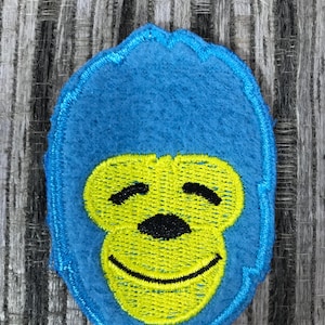 May include: A blue felt applique in the shape of a smiling gorilla face. The gorilla's face is yellow with black details, and the applique is outlined in blue stitching.