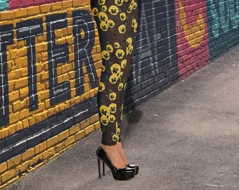 Op de afbeelding: Zwarte leggings met een bloemenpatroon van gele viooltjes op een donkerbruine achtergrond. De leggings worden gedragen door een persoon die voor een bakstenen muur met kleurrijke graffiti staat.