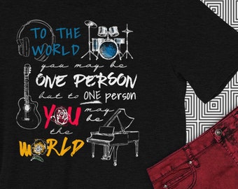 Op de afbeelding: Zwart gemarmerd T-shirt met een grafisch ontwerp met muziekinstrumenten, een roos en de tekst "To the world you may be one person but to one person you may be the world."