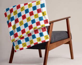 Op de afbeelding: Een plaid met een herhalend dambordpatroon van witte, rode, gele en blauwe vierkanten. Elk vierkant bevat een zwarte lijntekening van een boombox, een megafoon of een muzieknoot, wat zorgt voor een ritmisch en kleurrijk ontwerp.