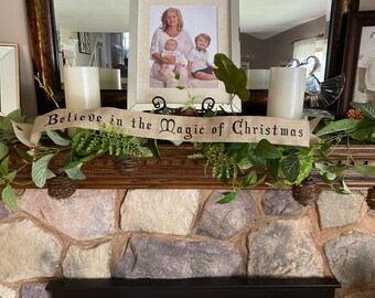 May include: A rustic fireplace mantel decorated with a green garland, two white candles, and a burlap banner that reads "Believe in the magic of Christmas".