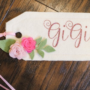 May include: A white felt tag with pink glitter lettering that reads "Gigi". The tag is decorated with pink felt flowers and green felt leaves. A pink ribbon is attached to the tag.