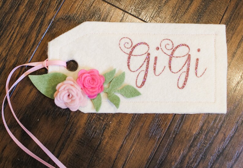 May include: A white felt tag with pink glitter lettering that reads "Gigi". The tag is decorated with pink felt flowers and green felt leaves. A pink ribbon is attached to the tag.