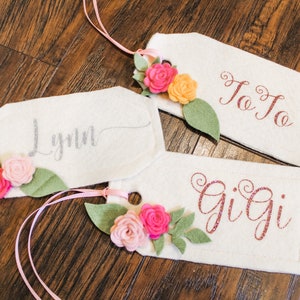 May include: Three white felt gift tags with pink, peach, and yellow felt flowers and green felt leaves. The tags are decorated with the names "Lynn", "Gigi", and "Jo Jo" in different colored embroidery.