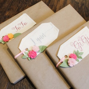 May include: Three brown gift boxes with white gift tags tied with pink ribbon. The tags are decorated with pink felt flowers and have the names "JoJo", "Lyla", and "Gigi" written on them.
