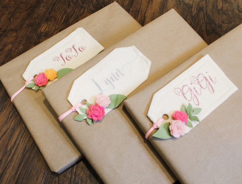 May include: Three brown gift boxes with white gift tags tied with pink ribbon. The tags are decorated with pink felt flowers and have the names "JoJo", "Lyla", and "Gigi" written on them.