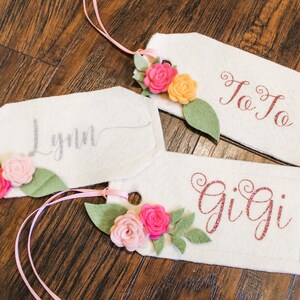 May include: Three white felt gift tags with pink, peach, and yellow felt flowers and green felt leaves. The tags are tied with pink ribbon and have the names Lynn, JoJo, and Gigi written on them in silver, red, and pink glitter.