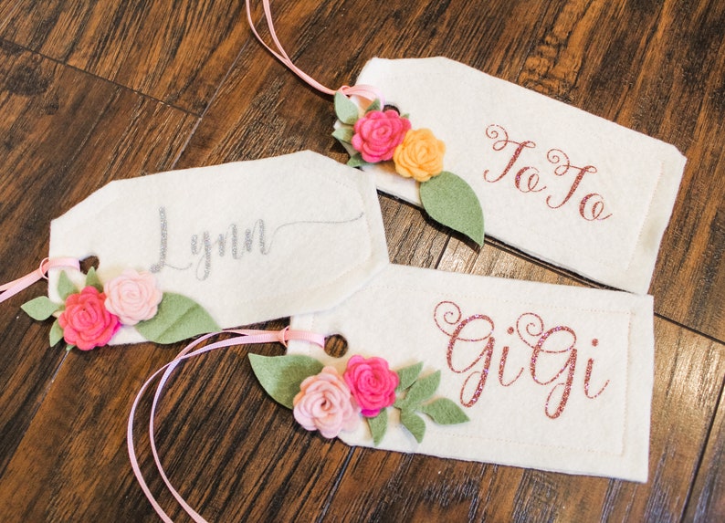 May include: Three white felt gift tags with pink, peach, and yellow felt flowers and green felt leaves. The tags are tied with pink ribbon and have the names Lynn, JoJo, and Gigi written on them in silver, red, and pink glitter.