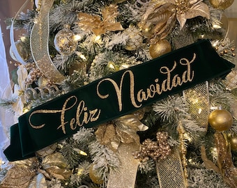 May include: A green velvet Christmas banner with gold glitter lettering that reads "Feliz Navidad" hanging on a Christmas tree decorated with gold and silver ornaments, ribbon, and lights.