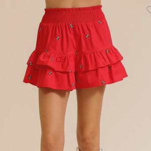 Könnte beinhalten: Rote Shorts mit einem gestuften Rüschendesign und Stickereien. Die Shorts haben einen gerafften Bund. Das Model trägt weiße Cowboy-Stiefel. Die Shorts sind ein auffälliges Kleidungsstück.