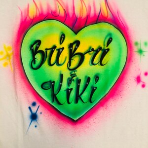 May include: A green heart with a pink outline and yellow flames behind it. The heart has the words "Bri Bri Kiki" written in black inside.