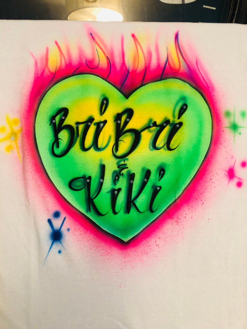 May include: A green heart with a pink outline and yellow flames behind it. The heart has the words "Bri Bri Kiki" written in black inside.