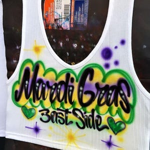 May include: White ribbed vest top with a scoop neck. The front displays airbrushed text reading "Mardi Gras Bast Side" in black, purple, green, and yellow, with heart and starburst designs.