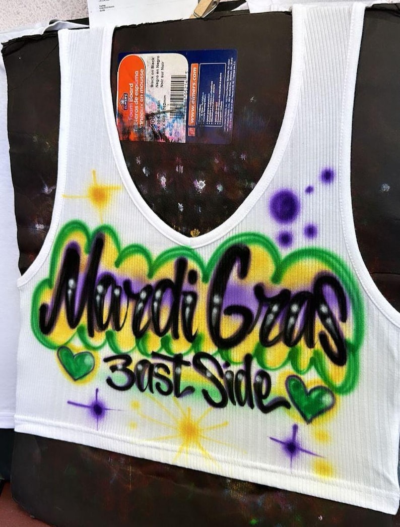 May include: White ribbed vest top with a scoop neck. The front displays airbrushed text reading "Mardi Gras Bast Side" in black, purple, green, and yellow, with heart and starburst designs.