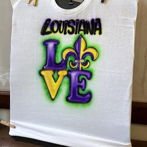 May include: White t-shirt featuring the words "LOUISIANA LOVE" in a purple, yellow, and green airbrushed design, including a fleur-de-lis symbol. The shirt is secured with wooden pegs.