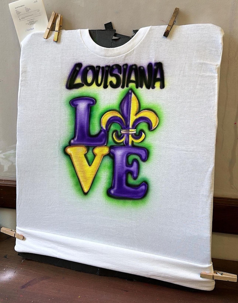 May include: White t-shirt featuring the words "LOUISIANA LOVE" in a purple, yellow, and green airbrushed design, including a fleur-de-lis symbol. The shirt is secured with wooden pegs.