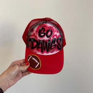 May include: Red trucker hat with the words "GO DAWGS" in black and white airbrushed on the front. A football patch is on the brim. The hat has a red mesh back.