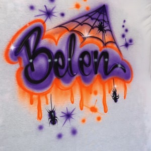 May include: White t-shirt with an airbrushed design featuring the name "Belen" in purple and black, outlined with orange. The design includes a spider web, spiders, and starbursts, creating a Halloween theme.