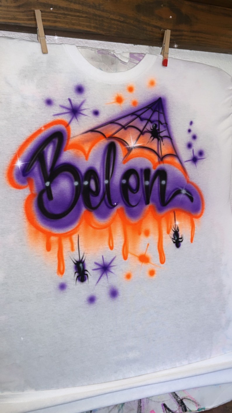 May include: White t-shirt with an airbrushed design featuring the name "Belen" in purple and black, outlined with orange. The design includes a spider web, spiders, and starbursts, creating a Halloween theme.