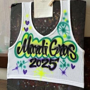 May include: White ribbed vest top with a V-neck, displaying the words "Mardi Gras 2025" in black graffiti-style lettering outlined in green and yellow. The design includes purple hearts and starbursts.