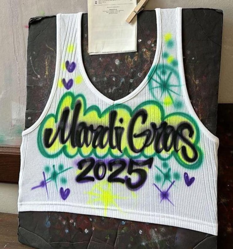 May include: White ribbed vest top with a V-neck, displaying the words "Mardi Gras 2025" in black graffiti-style lettering outlined in green and yellow. The design includes purple hearts and starbursts.