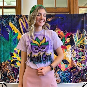 May include: A lilac t-shirt featuring a Mardi Gras mask design in purple, green, and yellow. The words "Mardi Gras" are written below the mask. The person is wearing a green beaded headband and a pink skirt.