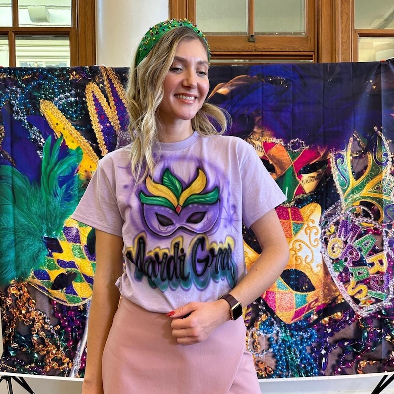 May include: A lilac t-shirt featuring a Mardi Gras mask design in purple, green, and yellow. The words "Mardi Gras" are written below the mask. The person is wearing a green beaded headband and a pink skirt.
