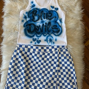May include: A white vest top and blue and white chequered skirt set. The vest top features the words "Blue Devils" in blue airbrush lettering with blue accents. The skirt is a wrap style with a blue and white checkerboard pattern.