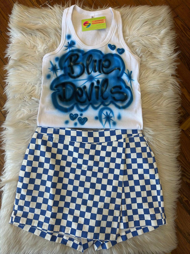 May include: A white vest top and blue and white chequered skirt set. The vest top features the words "Blue Devils" in blue airbrush lettering with blue accents. The skirt is a wrap style with a blue and white checkerboard pattern.