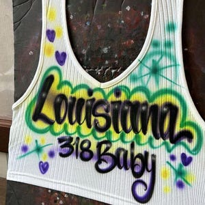 May include: White ribbed vest top with a V-neck, displaying the words "Louisiana 318 Baby" in a graffiti-style design. The design includes green, yellow, and purple accents, with heart and star details.