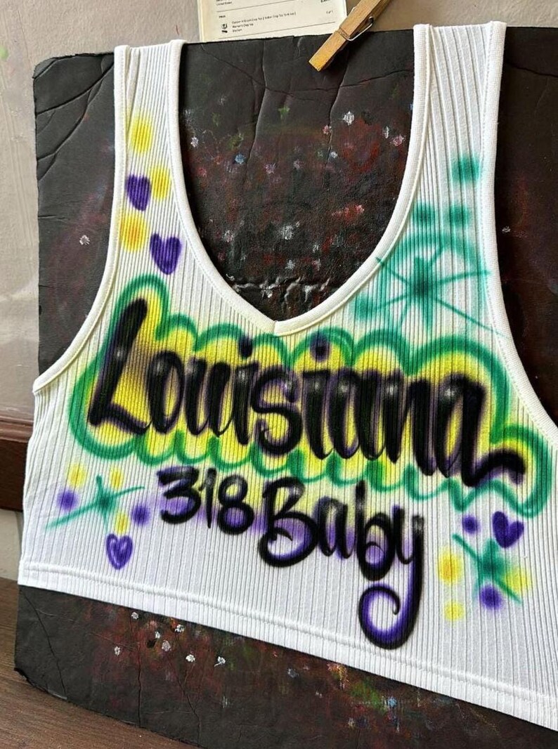 May include: White ribbed vest top with a V-neck, displaying the words "Louisiana 318 Baby" in a graffiti-style design. The design includes green, yellow, and purple accents, with heart and star details.