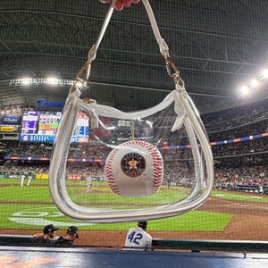 May include: A clear, transparent handbag with white trim and a white strap, holding a baseball with the Houston Astros logo. The bag is held up in front of a baseball stadium.