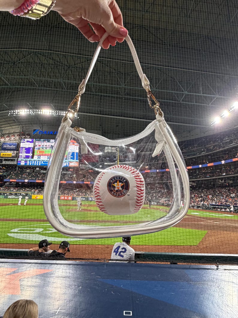 May include: A clear, transparent handbag with white trim and a white strap, holding a baseball with the Houston Astros logo. The bag is held up in front of a baseball stadium.