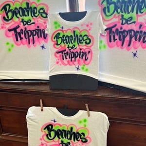 May include: White t-shirts with the phrase "Beaches be Trippin'" in a colorful, airbrushed design. The text is in black, outlined with pink and green accents. The shirts are displayed on a black mannequin and clothesline.
