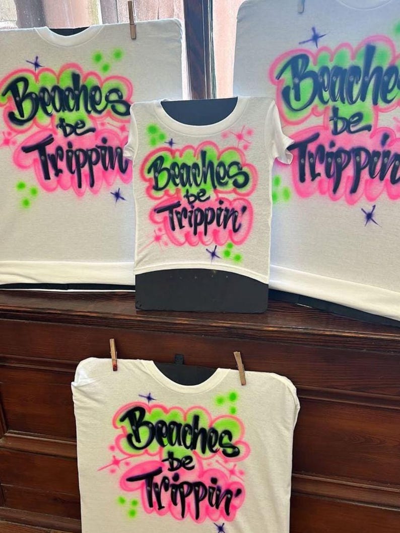 May include: White t-shirts with the phrase "Beaches be Trippin'" in a colorful, airbrushed design. The text is in black, outlined with pink and green accents. The shirts are displayed on a black mannequin and clothesline.