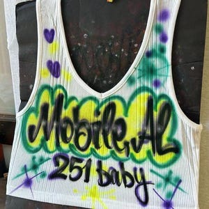 May include: White ribbed vest top with a V-neck, featuring graffiti-style text that reads "Mobile, AL 251 baby" in black, yellow, and green. Purple heart and star accents are also present.