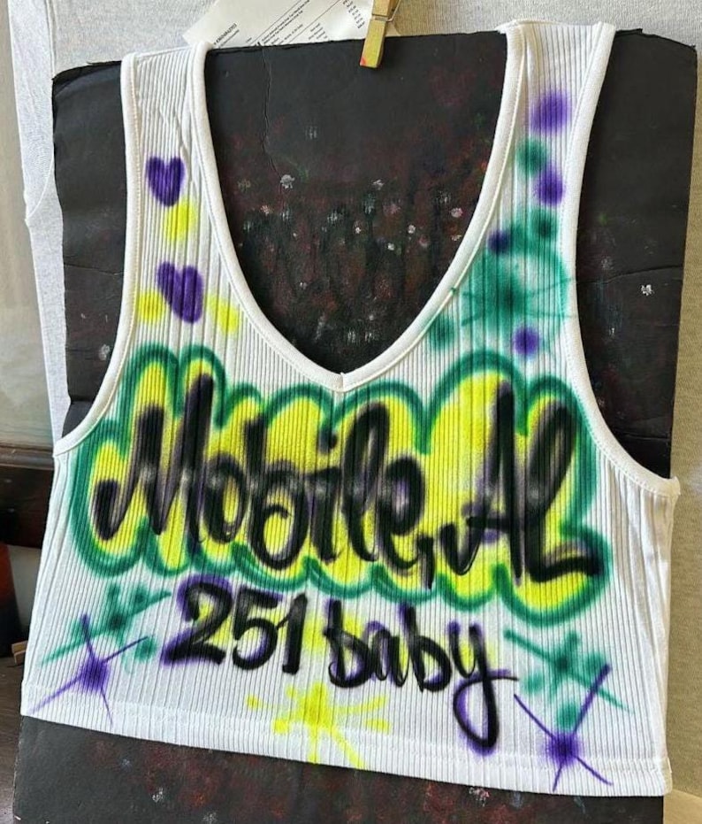 May include: White ribbed vest top with a V-neck, featuring graffiti-style text that reads "Mobile, AL 251 baby" in black, yellow, and green. Purple heart and star accents are also present.