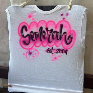 May include: White sleeveless t-shirt with the name "Semerah" in pink bubble letters, outlined in black, and the text "est. 2004" below. The shirt is held by clothespins.