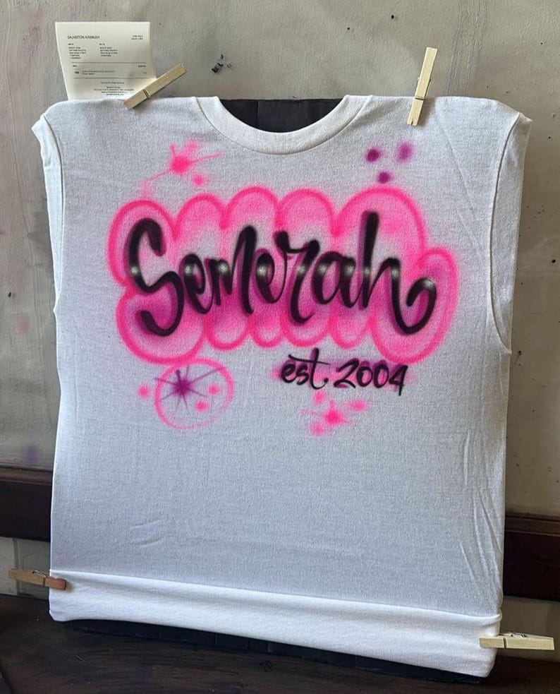 May include: White sleeveless t-shirt with the name "Semerah" in pink bubble letters, outlined in black, and the text "est. 2004" below. The shirt is held by clothespins.