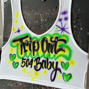 May include: White ribbed vest top with a V-neck. It has airbrushed text reading "Trip Out 504 Baby" in black and green, with purple accents and green heart graphics. A casual, sleeveless garment.