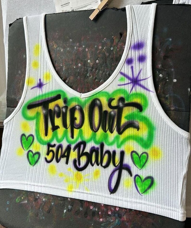 May include: White ribbed vest top with a V-neck. It has airbrushed text reading "Trip Out 504 Baby" in black and green, with purple accents and green heart graphics. A casual, sleeveless garment.