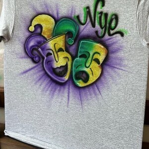 May include: A grey t-shirt with a vibrant airbrushed design. It features two theatre masks in yellow, green, and purple, with the word "Nye" in green script above. The masks are set against a purple burst.