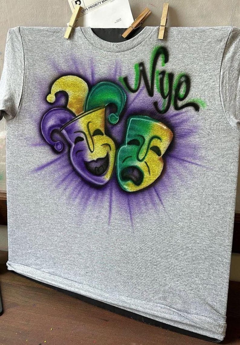 May include: A grey t-shirt with a vibrant airbrushed design. It features two theatre masks in yellow, green, and purple, with the word "Nye" in green script above. The masks are set against a purple burst.