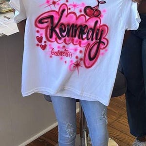 May include: White t-shirt with a pink and black airbrushed design. The design includes the name "Kennedy" in a cursive font, cherries, hearts, and the word "Galveston".