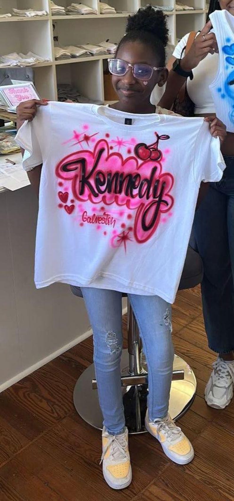 May include: White t-shirt with a pink and black airbrushed design. The design includes the name "Kennedy" in a cursive font, cherries, hearts, and the word "Galveston".
