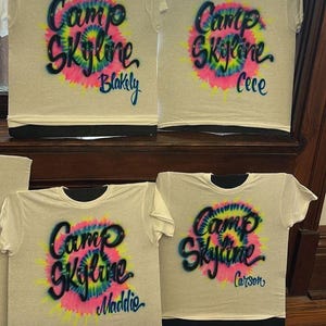 May include: Four white t-shirts with the words "Camp Skyline" in a tie-dye design. Each shirt has a different name: Blakely, Cece, Maddie, and Carson. The shirts have black collars and are displayed on a wooden surface.