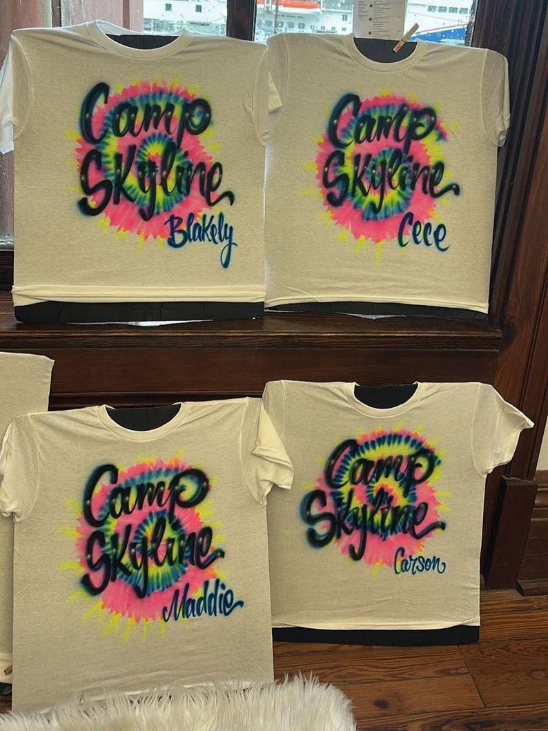 May include: Four white t-shirts with the words "Camp Skyline" in a tie-dye design. Each shirt has a different name: Blakely, Cece, Maddie, and Carson. The shirts have black collars and are displayed on a wooden surface.