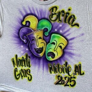 May include: A grey t-shirt with a Mardi Gras design. The design features two stylized masks in yellow, green, and purple, with the words "Bria", "Mardi Gras", "Mobile, AL", and "2025" in a colourful, airbrushed style.