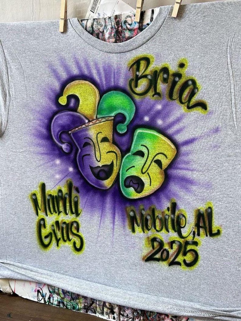 May include: A grey t-shirt with a Mardi Gras design. The design features two stylized masks in yellow, green, and purple, with the words "Bria", "Mardi Gras", "Mobile, AL", and "2025" in a colourful, airbrushed style.