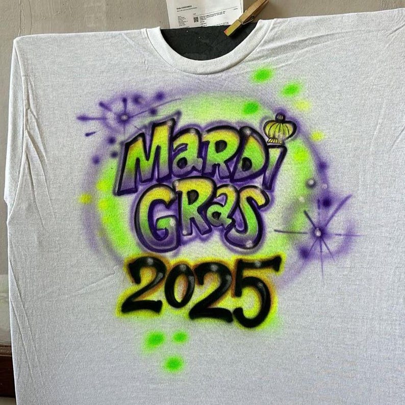 May include: A white t-shirt with a vibrant airbrushed design. The design features the words "Mardi Gras 2025" in green and purple, with a yellow crown. The shirt has a crew neck.